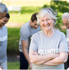Older woman wearing a t-shirt which reads volunteer. She is smiling to camera and a group of volunteers are working together behind her.