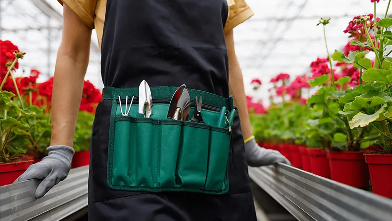 A woman wearing a garden apron filled with garden tools.