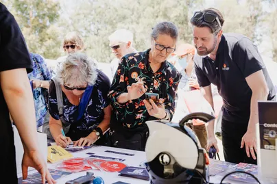 Image of people looking at different products at a stall. A woman is showing something on her phone to the man from the stall.