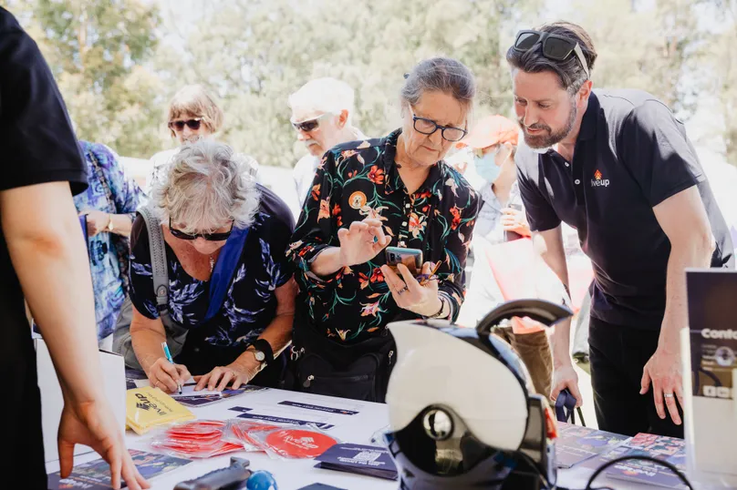 Image of people looking at different products at a stall. A woman is showing something on her phone to the man from the stall.