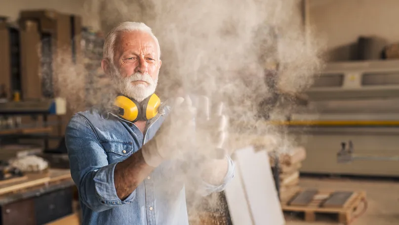 Man with ear protection and gloves working in a woodshop. Sawdust is flying in the air.