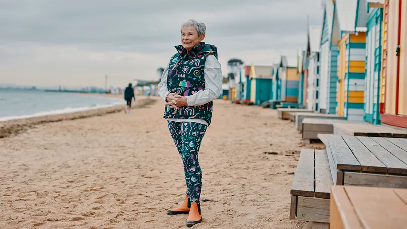 An older woman smiling at the beach. She is dressed in colourful warm clothes, on an overcast day. 