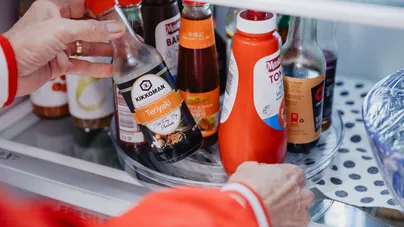 Hands turning a lazy suzan around to reach various sauces from a crowded fridge shelf.