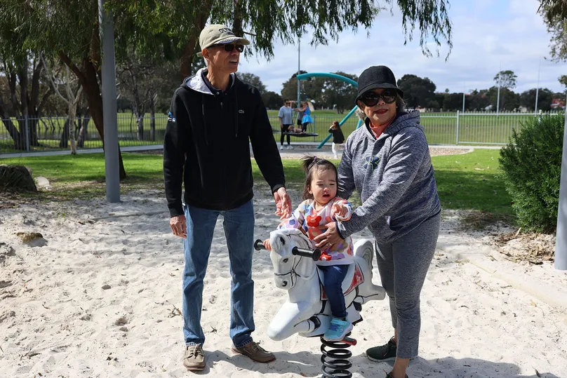 Two grandparents play with their granddaughter on the swinging horse at the park.