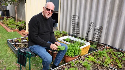 An older man sitting on a garden kneeler in his veggie garden.