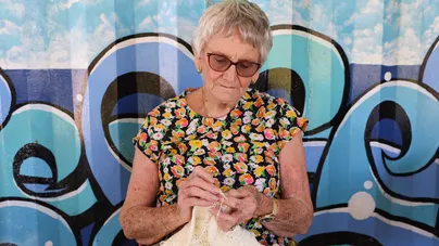 Image of an older woman knitting outside, standing in front of a colourfully painted wall.