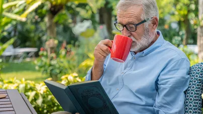 An older man in glasses, drinking tea while reading a book in the park.