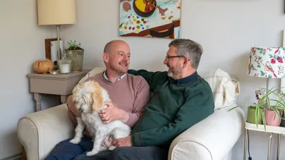 Two men sit on an armchair together with their dog. One man has his arm around the other's shoulder as they smile at each other.