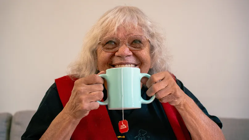 Image of older woman drinking tea out of a two-handled mug. She is holding both handles and smiling.