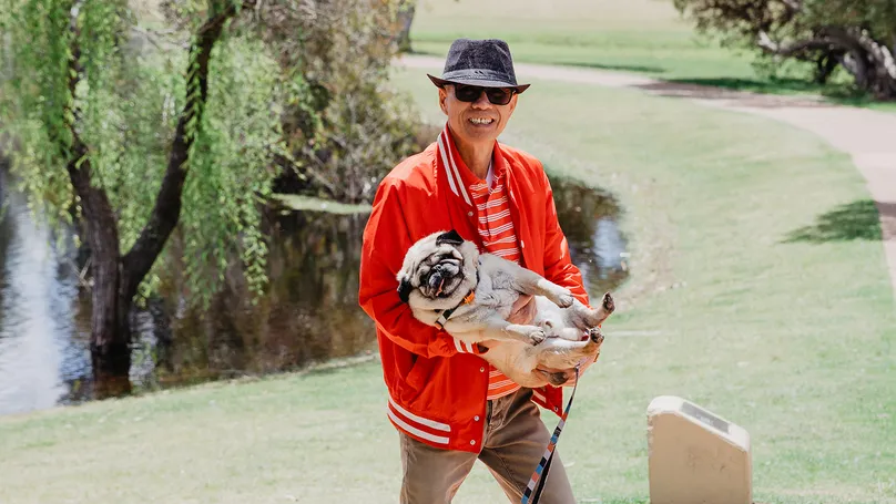 Image of a man walking through a park. He is holding a happy pug in his arms.