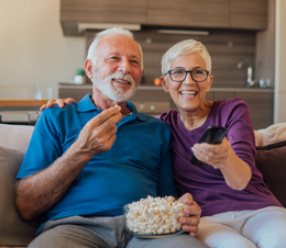 An older couple sitting on the couch watching TV together. They hold popcorn and the TV remote control in their hands.